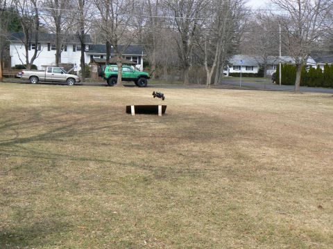 1994 Ford Bronco - An RC car flies through the air over a ramp in a grassy yard with two full-size vehicles parked nearby.