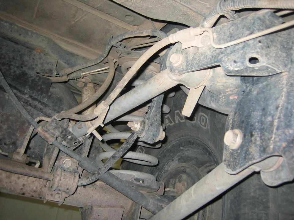 Underside view of a vehicle's rear suspension with coils, shocks, and brake lines visible. Tire tread is partially visible