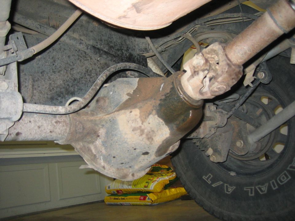 Driveshaft connected to a rusty differential, with a tire and exhaust pipe visible