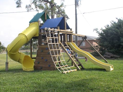 1993 Ford C-350 (Centurion Conversions) - A backyard playset features a yellow tube slide, a wavy slide, and a rope climbing wall, set against a backdrop of trees and a house.