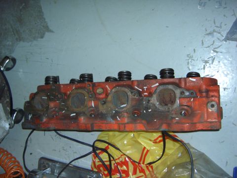 1974 Ford Bronco - An orange engine cylinder head with valve springs and exhaust ports is shown in a workshop setting.