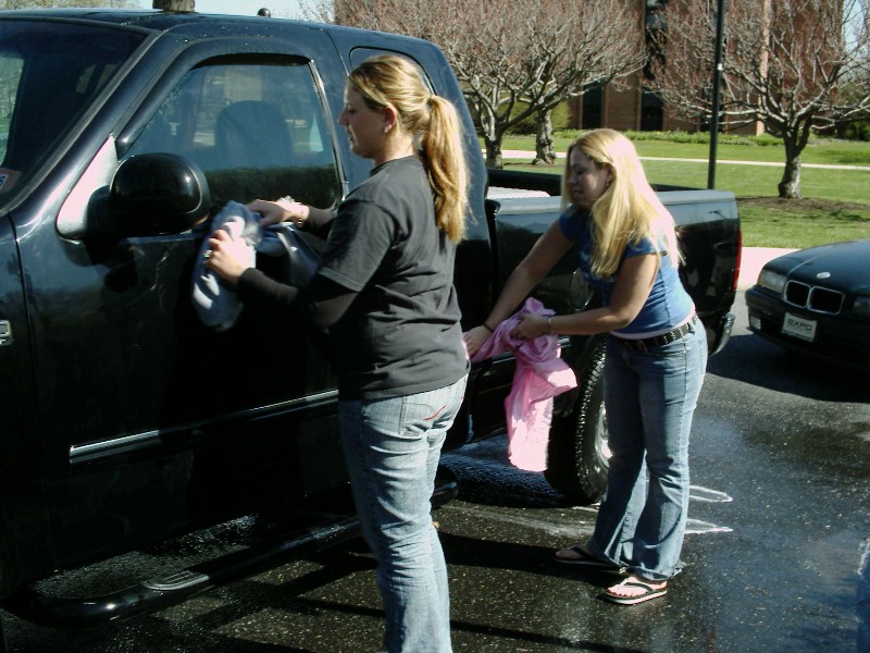 Two women washing a black pickup truck. Another car is parked nearby