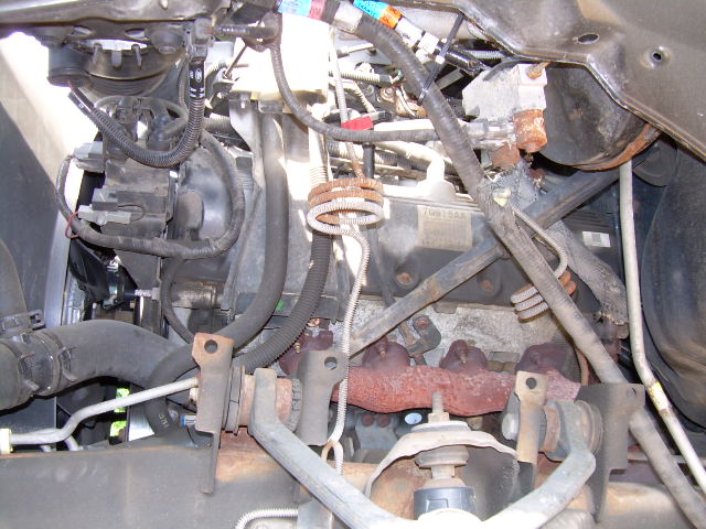 Engine bay view showing exhaust manifold, hoses, and wiring. Some rust is visible on the manifold