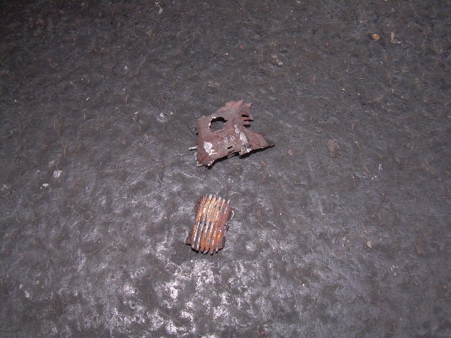 Two rusted metal pieces, one flat and jagged, the other cylindrical and ribbed, lie on a dark, textured surface
