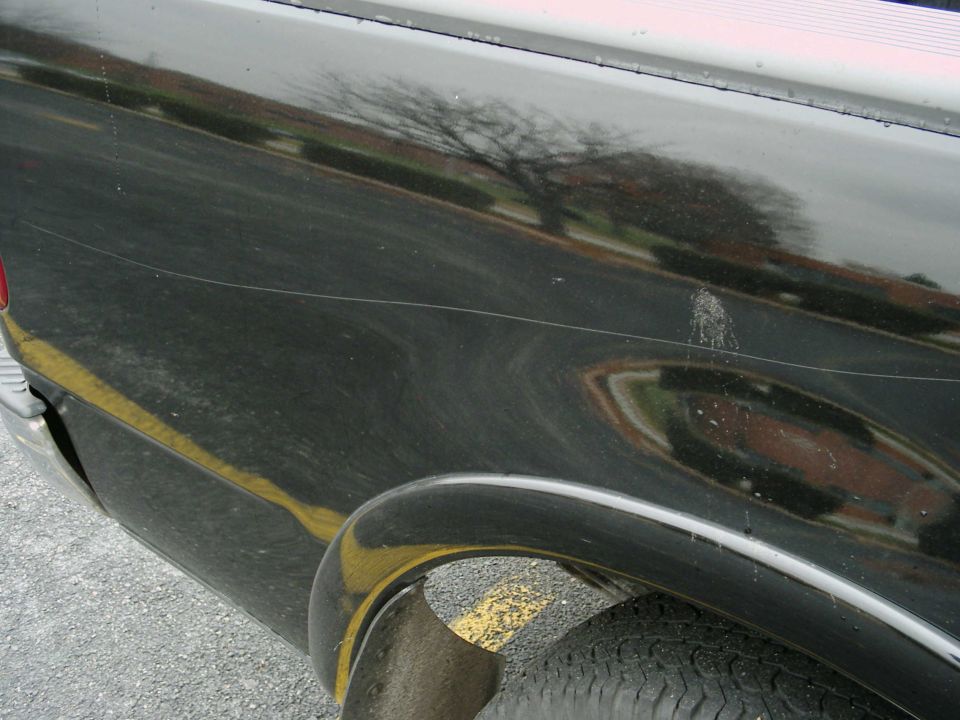 Black truck side panel with scratches and a yellow line, showing a reflection of a tree