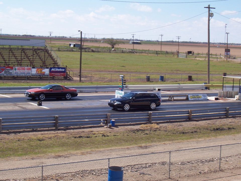 Two cars, a red coupe and a black wagon, race down a drag strip with bleachers and fields in the background