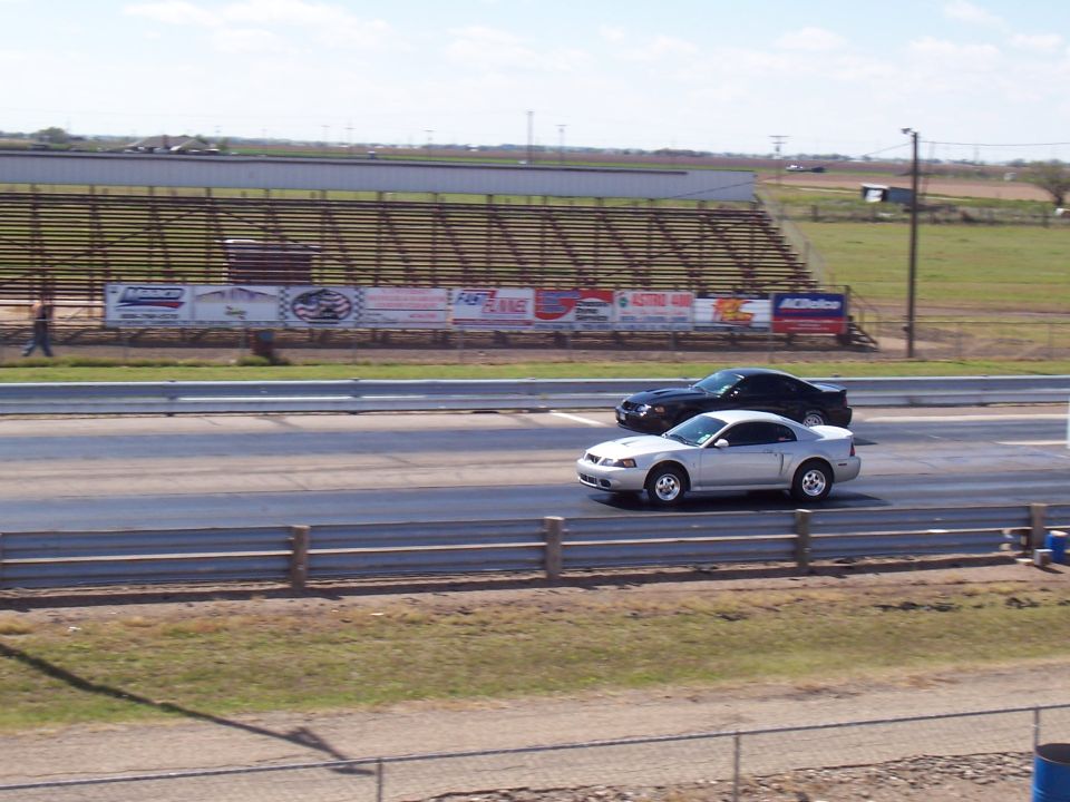 Silver Ford Mustang and black car racing on a drag strip with bleachers in the background