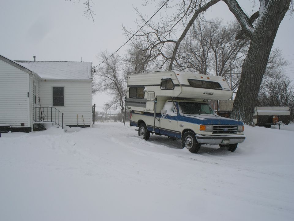 White and blue Ford pickup with cab-over camper parked in snow next to a white house