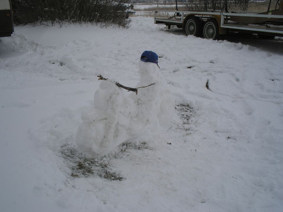 Snowman with a blue cap and stick arms in a snowy yard with a trailer in the background
