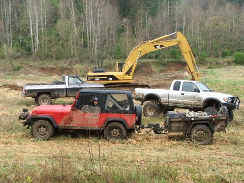1996 Ford Bronco - A red Jeep Wrangler tows a trailer through a muddy field, accompanied by a white pickup truck and a large yellow excavator.