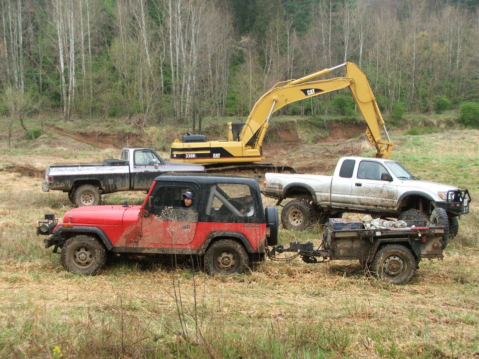 Red Jeep Wrangler towing a trailer, with a white pickup and yellow excavator behind it