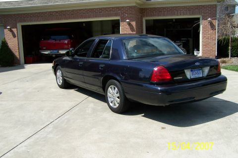 2003 Ford Crown Victoria - Rear view of a dark blue 2003 Ford Crown Victoria parked outside a brick garage with a red truck inside.