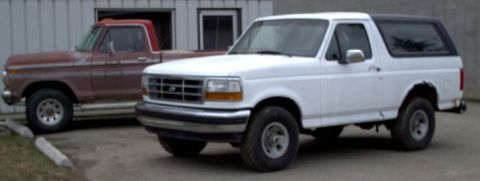 1990 Ford Bronco - A white Ford Bronco and a red Ford pickup truck are parked side-by-side in front of a building.