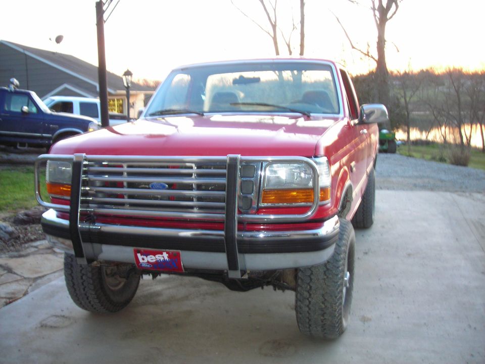Front view of a red 1995 Ford F-250 with a chrome grille guard. Another blue truck is parked behind it