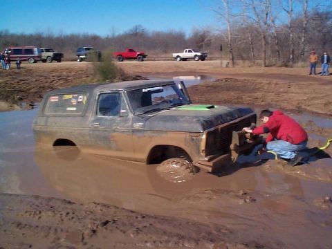 1978 Ford Bronco - A person works on a stuck 1978 Ford Bronco in a muddy off-road area, with other vehicles visible in the background.