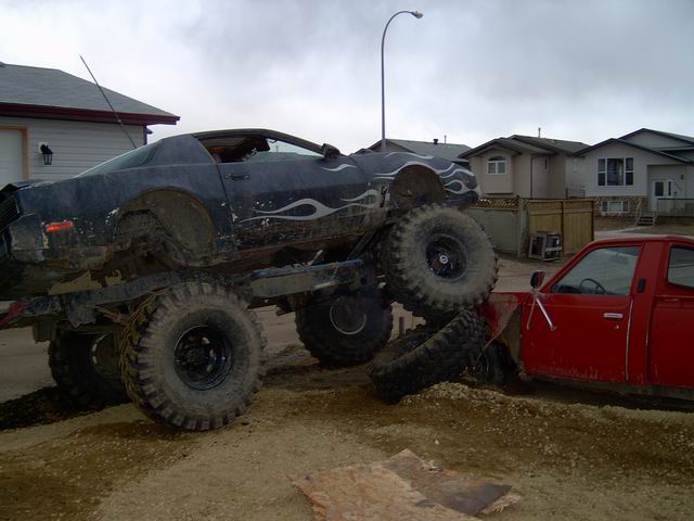 Dark monster truck with flame decals on top of a red pickup truck, tires are muddy