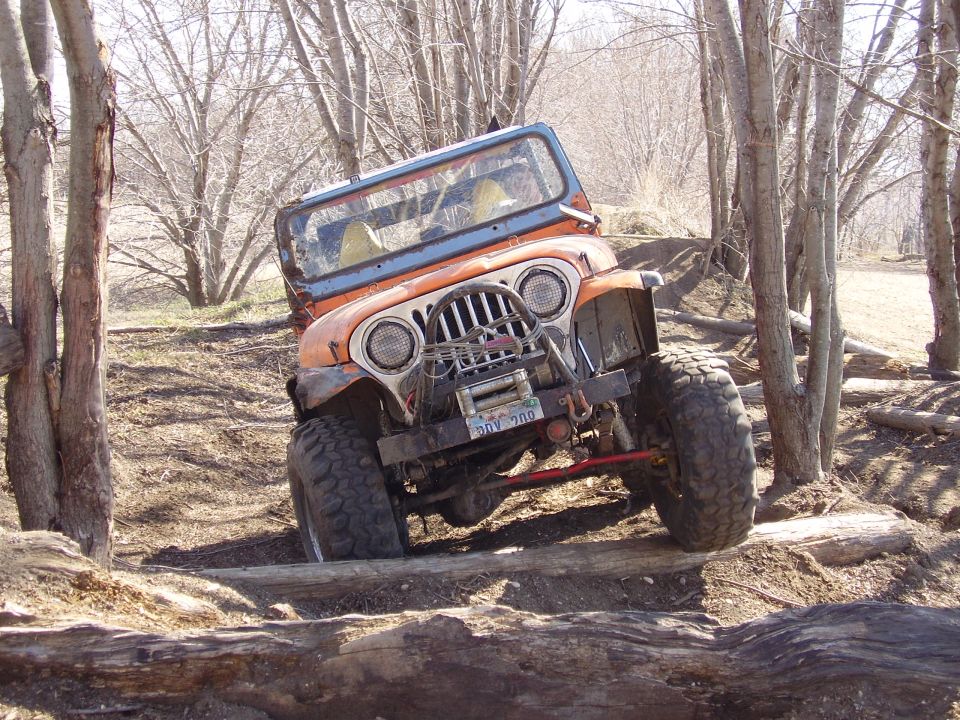 Orange Jeep CJ crawling over logs on a wooded off-road trail, front view