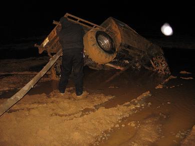 2005 Ford Excursion - A person stands near a heavily mud-covered off-road vehicle that has tipped onto its side in a muddy area at night.