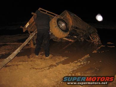 1987 Suzuki Samurai - An off-road vehicle has flipped over in the mud at night, with a person standing nearby.