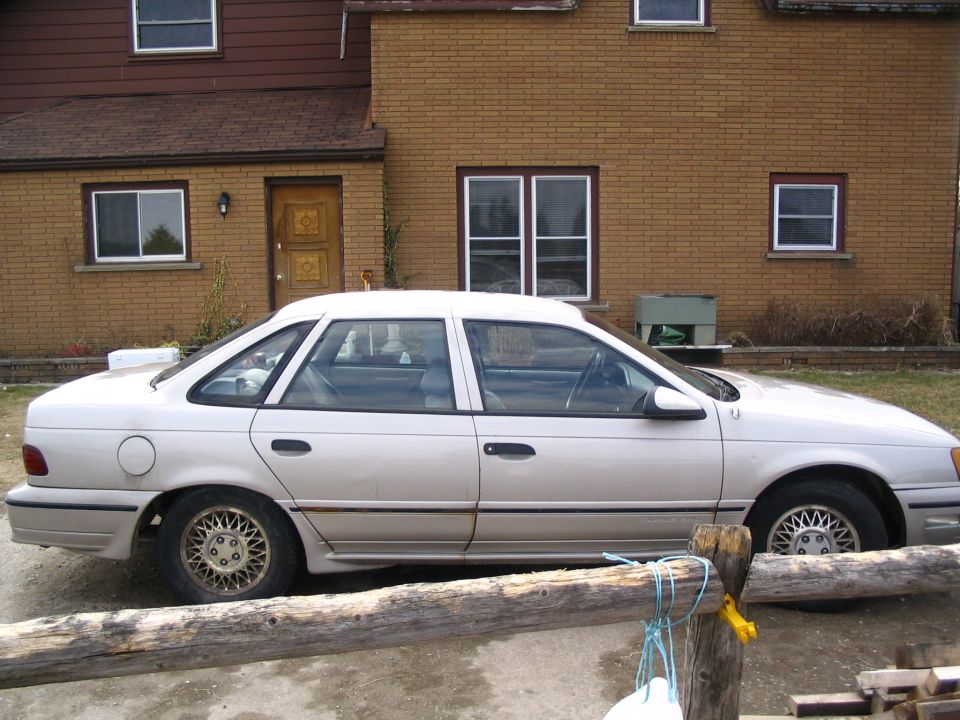 Light-colored 1989 Ford Taurus sedan parked in front of a brick house, side view