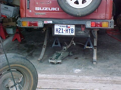 1978 Ford Bronco - The rear of a red Suzuki Samurai is shown elevated on jack stands, with a floor jack visible underneath.