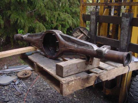 1979 Ford Bronco - A rusty rear axle housing is mounted on a wooden pallet outdoors, with trees and industrial equipment in the background.