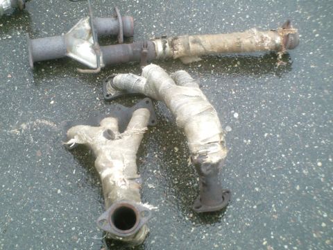 1995 Ford Probe - Two exhaust manifold sections and a pipe, all wrapped in heat-resistant material, lie on a wet surface.