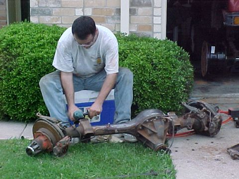 1978 Ford Bronco - Man works on a vehicle axle assembly outdoors, with bushes and a building in the background.