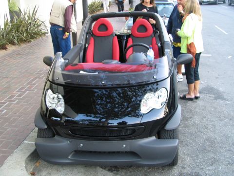 1986 Pontiac Fiero - A black Smart Fortwo Cabriolet with distinctive red seats is parked curbside, with several people observing it.