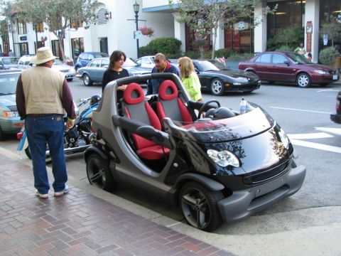 1986 Pontiac Fiero - A small, two-seater convertible is parked curbside with people standing around it.