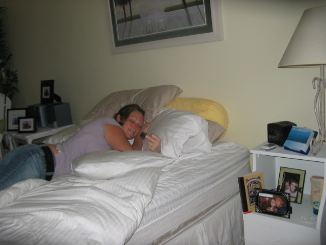 Woman in purple shirt and jeans lies on a white bed, holding a small object. Pillows and a lamp are visible