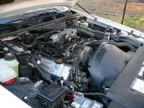 1997 Mercury Grand Marquis - Engine compartment of a white vehicle with V-configuration engine, alternator, intake ducting and battery.