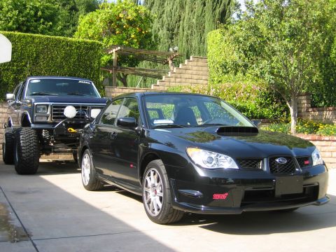 2007 Subaru Impreza - A black 2007 Subaru Impreza WRX STI sedan is parked in a driveway, with a lifted pickup truck behind it.