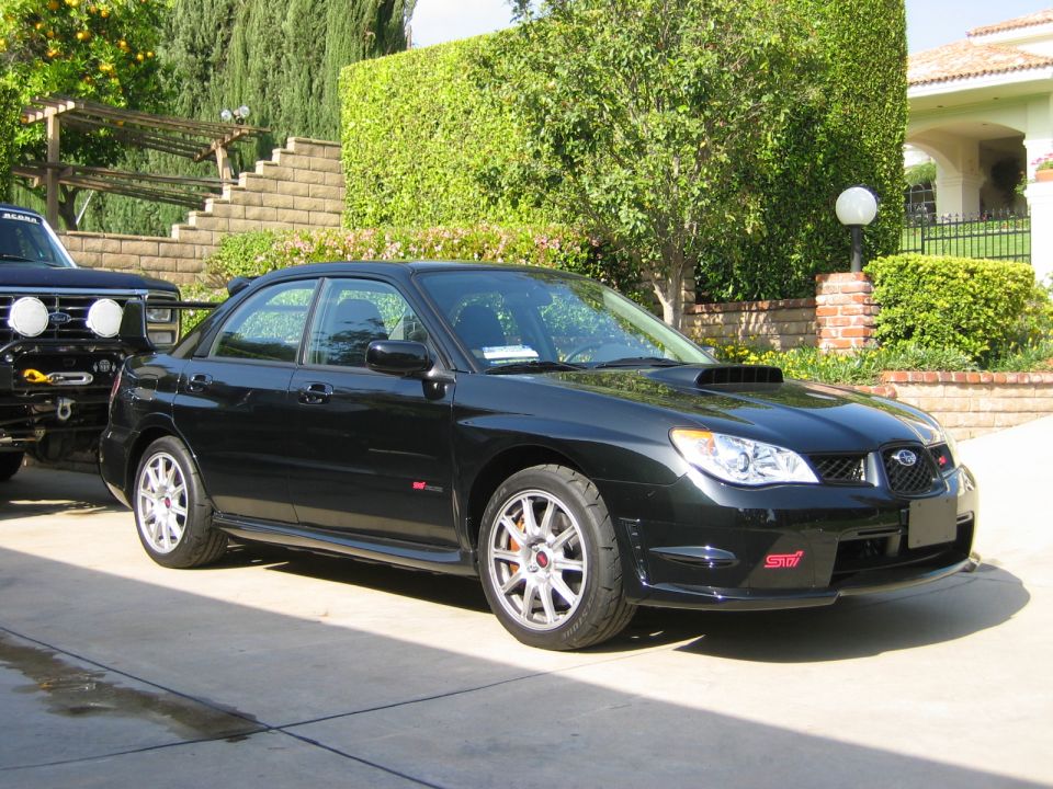 Black Subaru Impreza WRX STI sedan with a hood scoop and red STI badges, parked next to a dark SUV