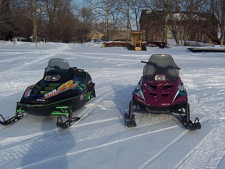 1996 Arctic Cat Puma - Two snowmobiles, one a 1996 Arctic Cat Puma, sit on a snowy track.