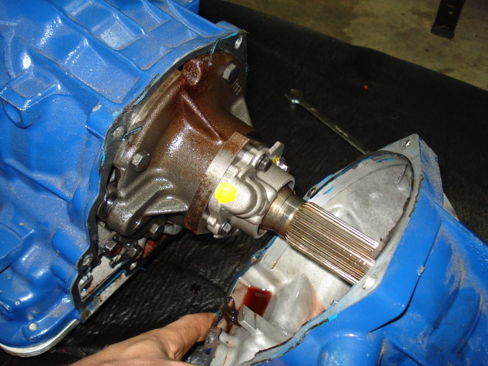 Blue transmission housing with rusty metal components and a splined shaft. A hand points to red fluid