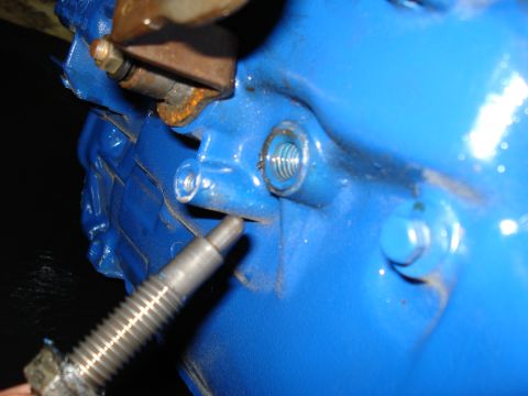 1984 Ford Bronco - Close-up of a blue painted engine component, showing a bolt and a threaded hole. Text at bottom reads "SUPERMOTORS.NET".