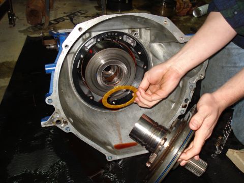 1984 Ford Bronco - A mechanic works on a transmission, holding a yellow gasket near the torque converter and housing.