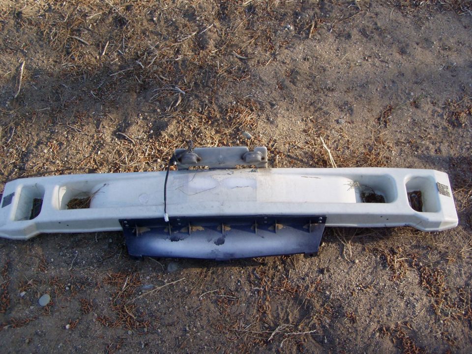 White bumper with dark lower valance lying on dirt ground, removed from vehicle