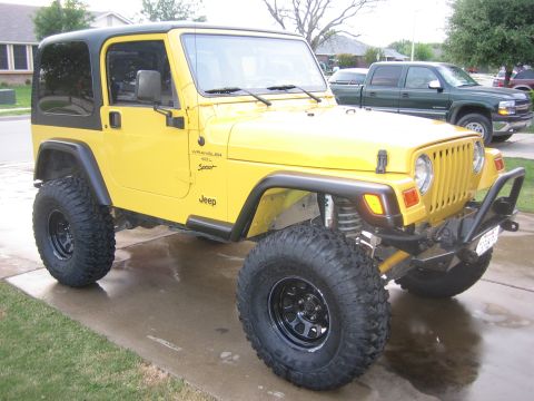2001 Jeep Wrangler - Passenger-side front three-quarter view of a lifted yellow Jeep Wrangler Sport with 4.0L badging, parked on a wet suburban driveway. It has…