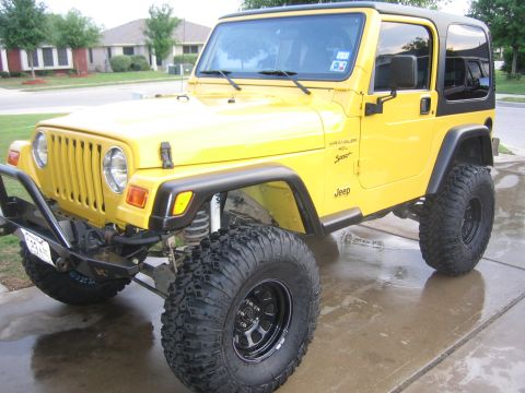 2001 Jeep Wrangler - Front three-quarter driver-side view of a lifted yellow Jeep Wrangler TJ with black hardtop, parked on a wet suburban driveway. Visible det…