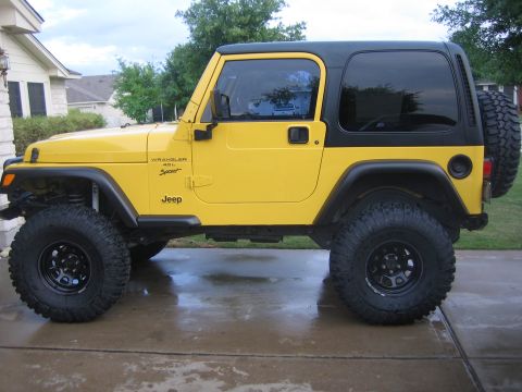 2001 Jeep Wrangler - Driver-side profile of a lifted yellow Jeep Wrangler TJ with a black hardtop, parked on a wet concrete driveway in front of a stone house.…