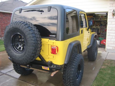 2001 Jeep Wrangler - Rear three-quarter view of a lifted yellow Jeep Wrangler TJ with a black hardtop on a wet driveway in front of an open home garage. A full-…