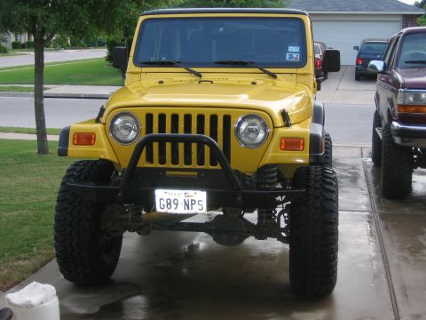 2001 Jeep Wrangler - Head-on front view of a lifted yellow Jeep Wrangler TJ on a wet suburban driveway, showing the seven-slot grille, round headlights, black t…