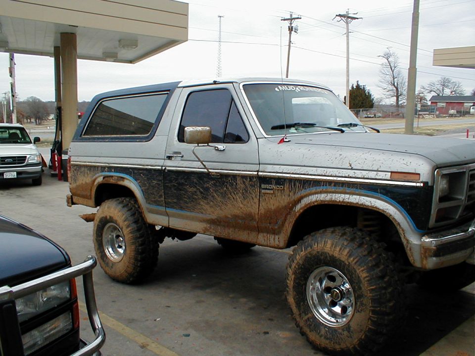 Side view of lifted silver/blue Ford Bronco on mud tires, parked at gas station