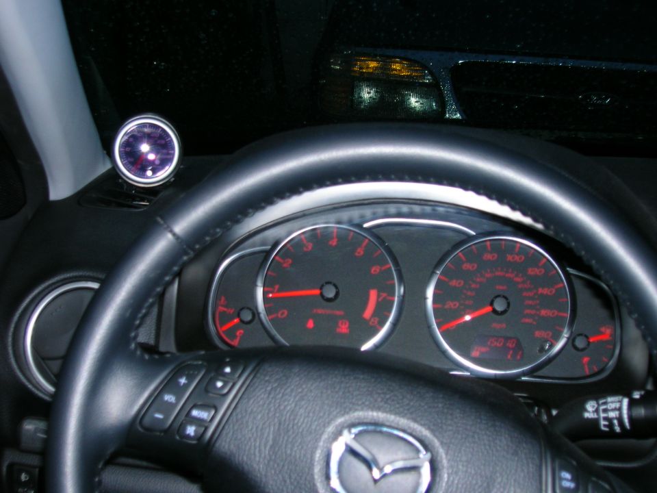 Car interior showing dashboard with speedometer, tachometer, and a silver-rimmed boost gauge. Steering wheel has audio controls. Mazda logo visible on steering wheel