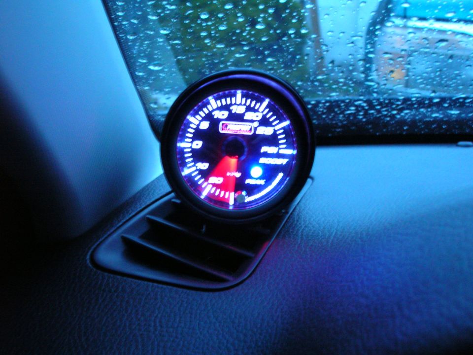 Interior view of a car dashboard with a glowing blue boost gauge mounted above an air vent. Water droplets are visible on the windshield