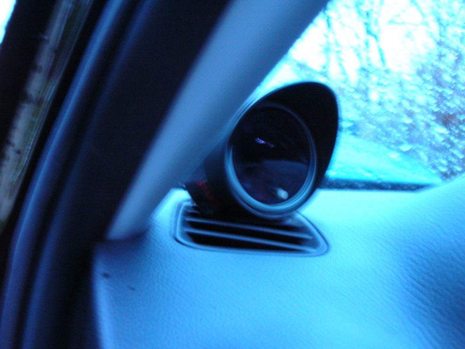 Aftermarket gauge installed on dashboard near air vent, with a blurry view of trees through the windshield