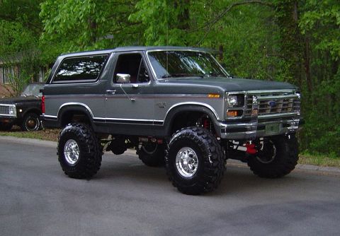 1986 Ford Bronco - A lifted gray two-door Ford Bronco SUV shown from a 3/4 front angle, sitting on large chrome wheels with oversized off-road mud tires and a…