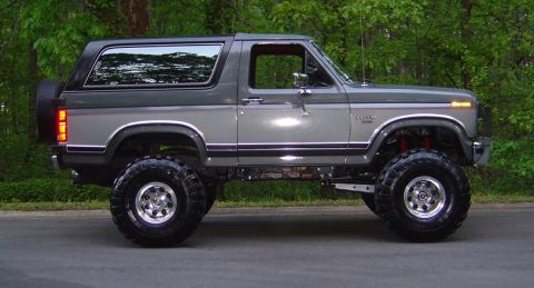 1986 Ford Bronco - A two-tone gray and silver 1980s Ford Bronco full-size SUV shown in side profile, lifted on large off-road tires with polished alloy wheels…